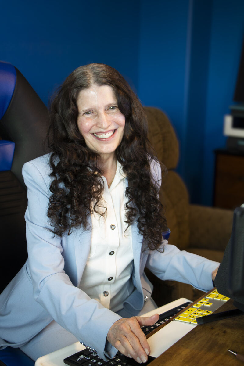 Victoria Essner sits smiling at her workstation, wearing a light blue blazer and white blouse, with a large-print yellow-and-black keyboard and computer equipment in front of her. Victoria Essner sits smiling at her workstation, wearing a light blue blazer and white blouse, with a large-print yellow-and-black keyboard and computer equipment in front of her.