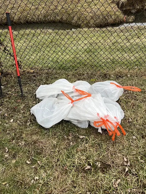 Bagged pet waste and a cleanup rake beside a chain-link fence.
