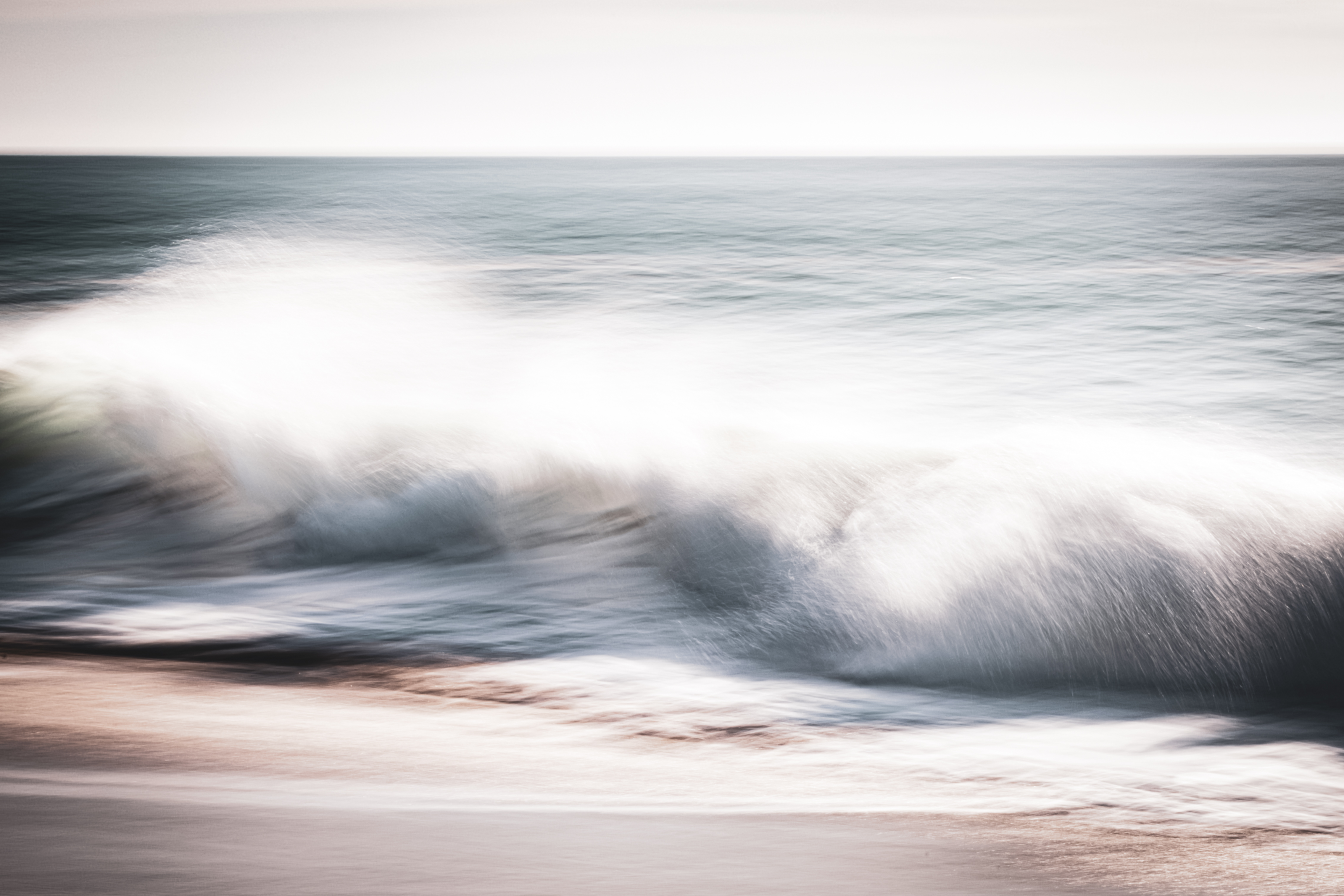 Long exposure ocean photograph of a softly breaking wave at Huntington Beach, California — pale blue and white tones with gentle motion blur. The final image in the Coastal Blur tribute series by Angie Harker. "All the Way to Shore."
