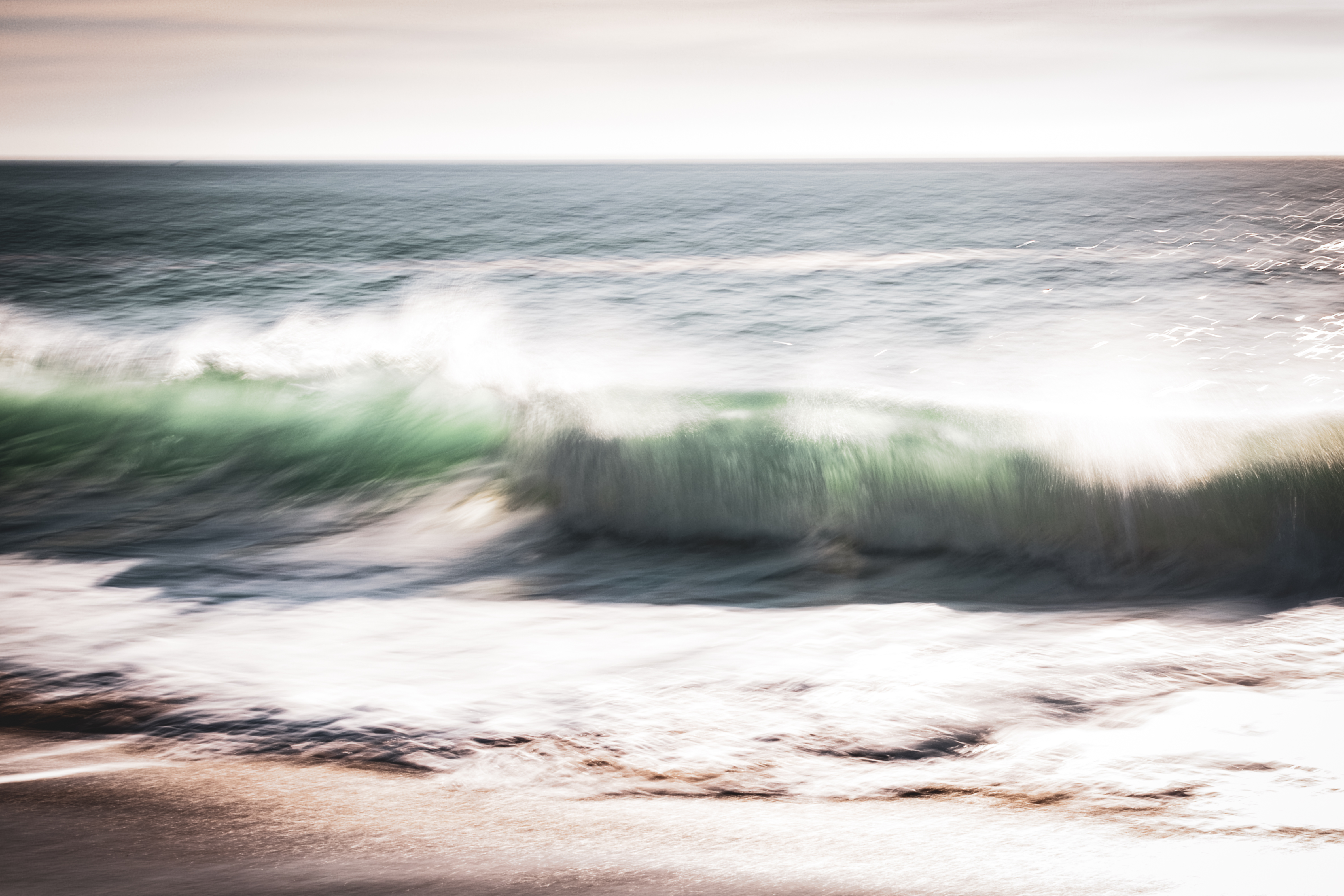 Long exposure ocean photograph of a breaking wave at Huntington Beach, California — soft green water cresting into white foam, captured with intentional camera movement. Part of the Coastal Blur series by Angie Harker. "What the Light Carried In."