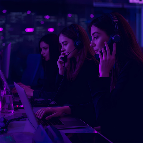 Three women wearing headsets working at computers in a dimly lit call center with purple ambient lighting.