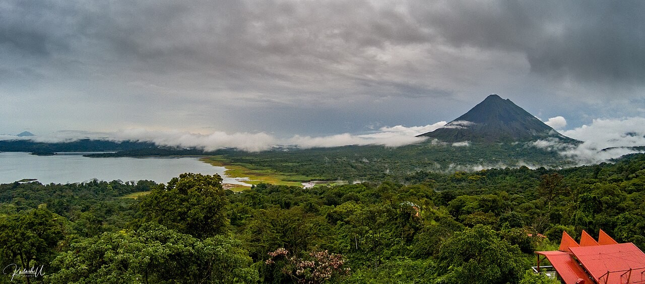 Arenal volcano, lake and forest beneath low clouds, with a red roof at the edge.
