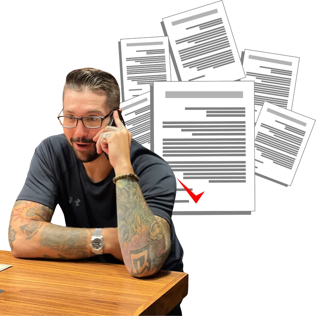 Drewbie Wilson on a phone call, leaning on a wooden table in glasses and a dark t-shirt, with a spread of document pages and a red check mark behind him