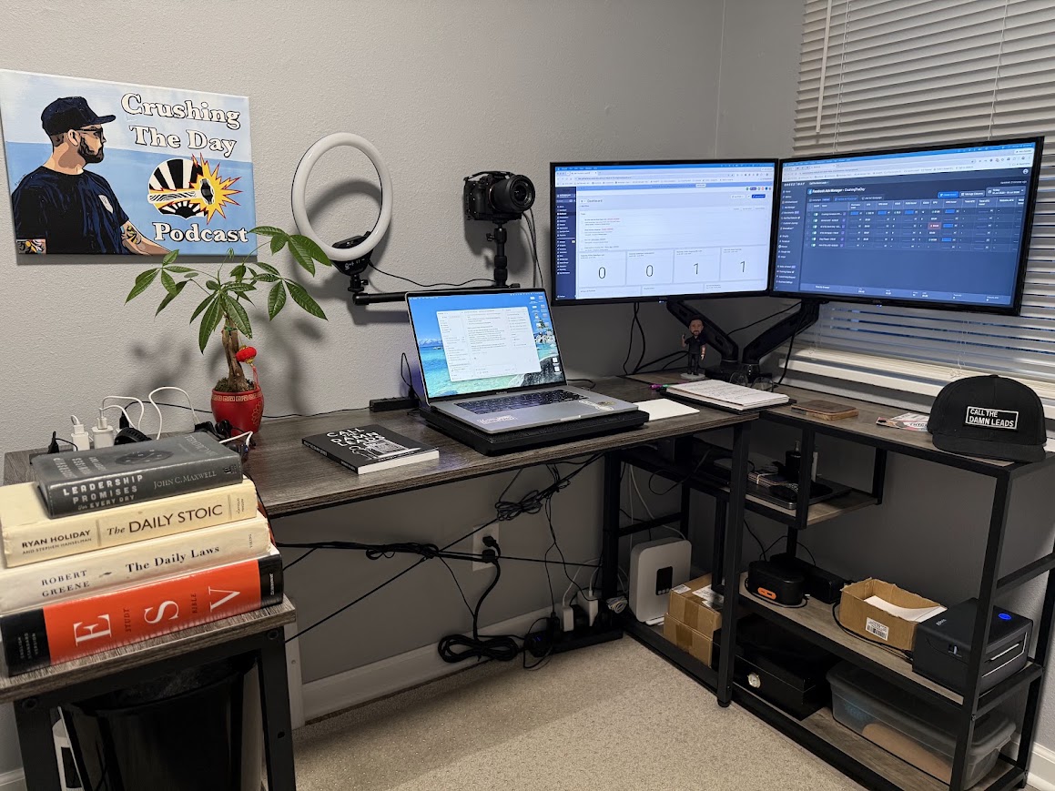 Drewbie Wilson's desk with two monitors, a laptop, the Call The Damn Leads book and hat, and Crushing The Day podcast art on the wall