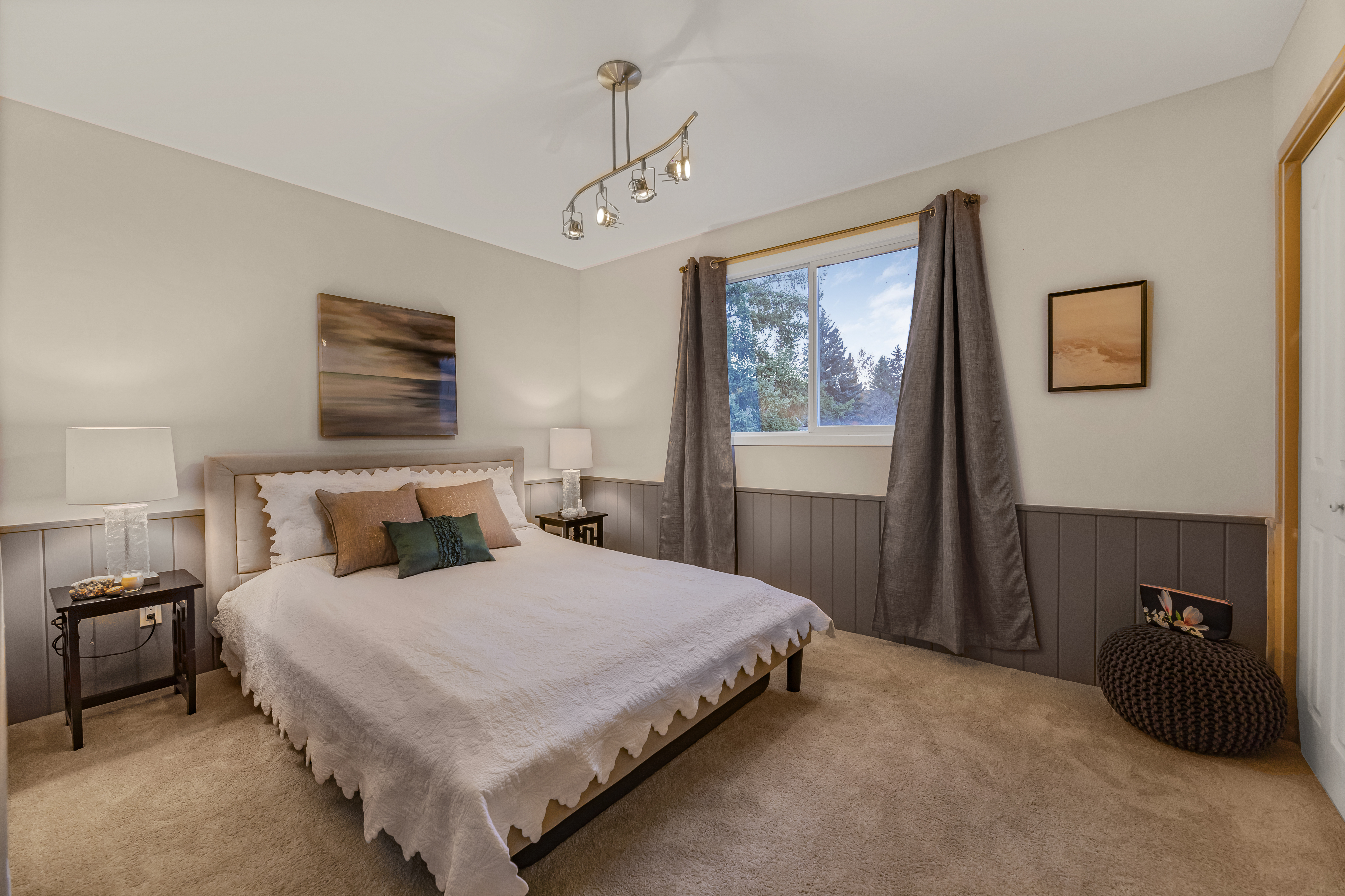 Staged bedroom with neutral bedding, accent pillows, wall art, and a large window with curtains letting in natural light.