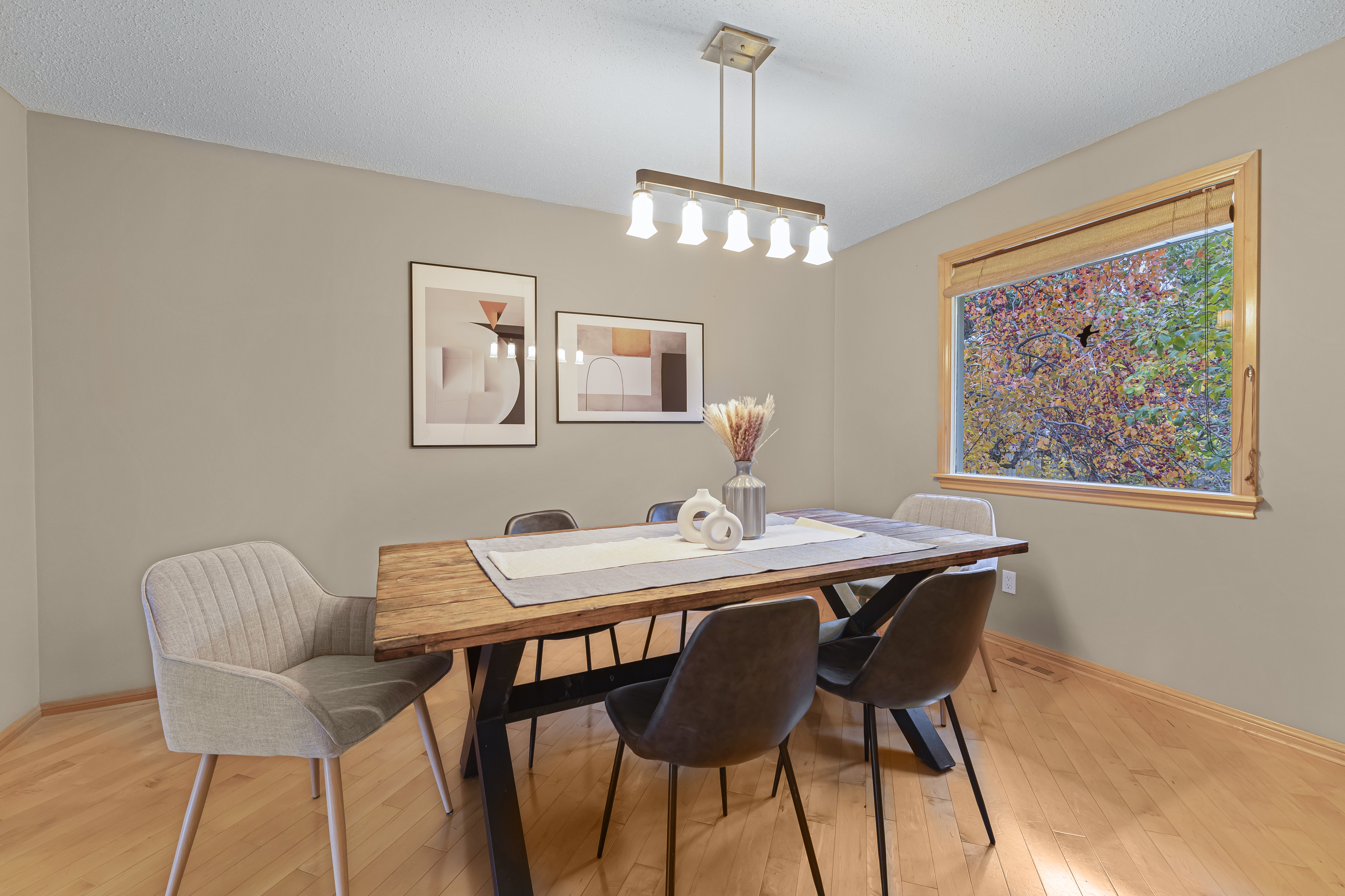 Staged dining room with a wood table, mixed seating, modern light fixture, framed wall art, and a window bringing in natural light.