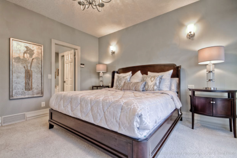 Bright staged bedroom with a wooden bed frame, neutral bedding, decorative pillows, wall sconces, and a bedside lamp.