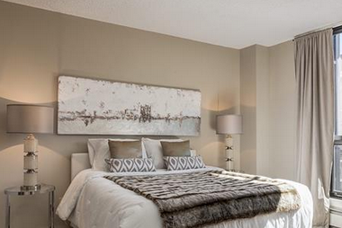Staged bedroom with a neatly made bed, patterned accent pillows and throw, large abstract artwork above the headboard, and a bedside lamp against soft neutral walls.