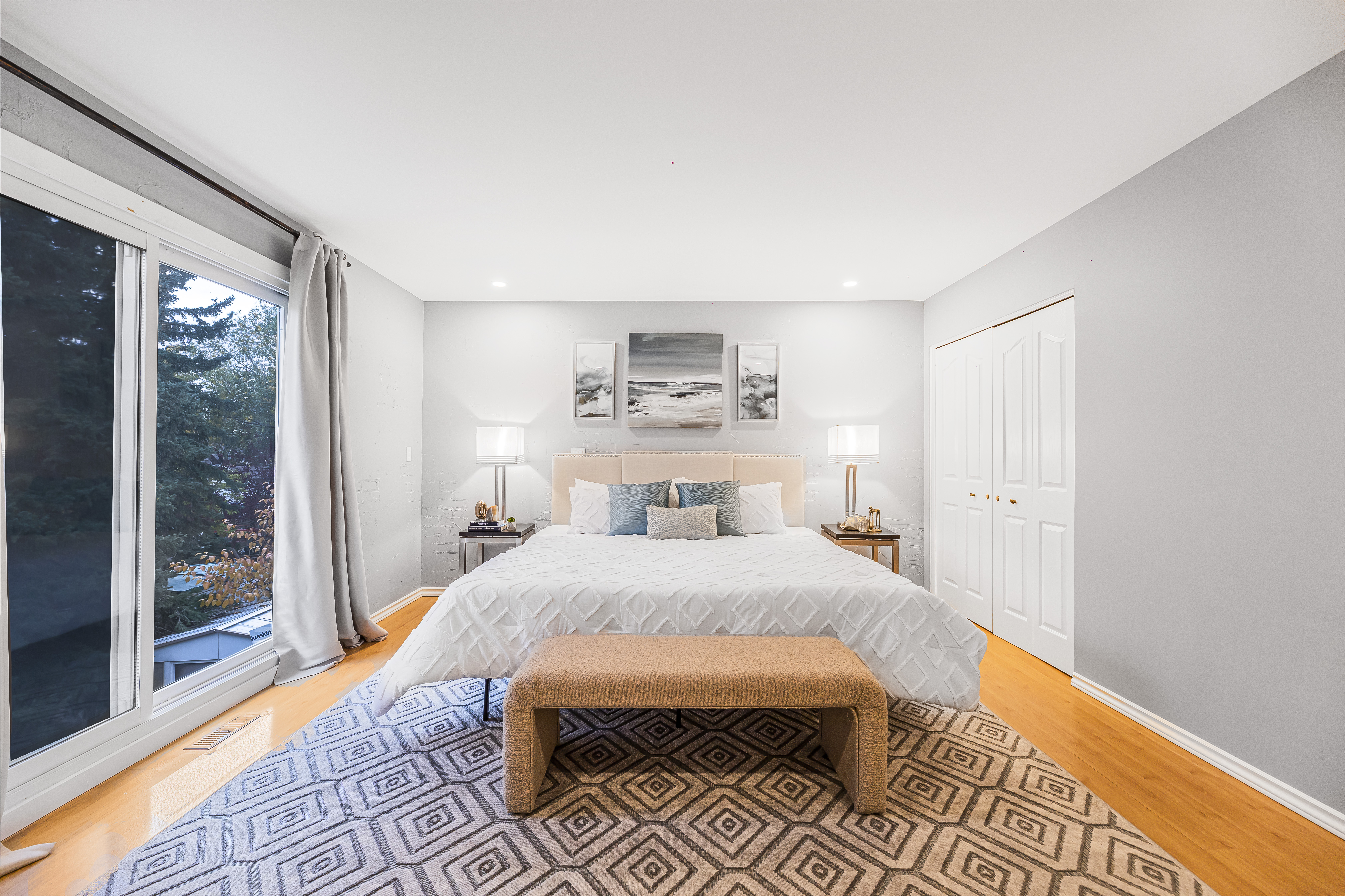 Staged bedroom with a large bed, neutral bedding and accent pillows, bench at the foot of the bed, and matching nightstands with lamps.