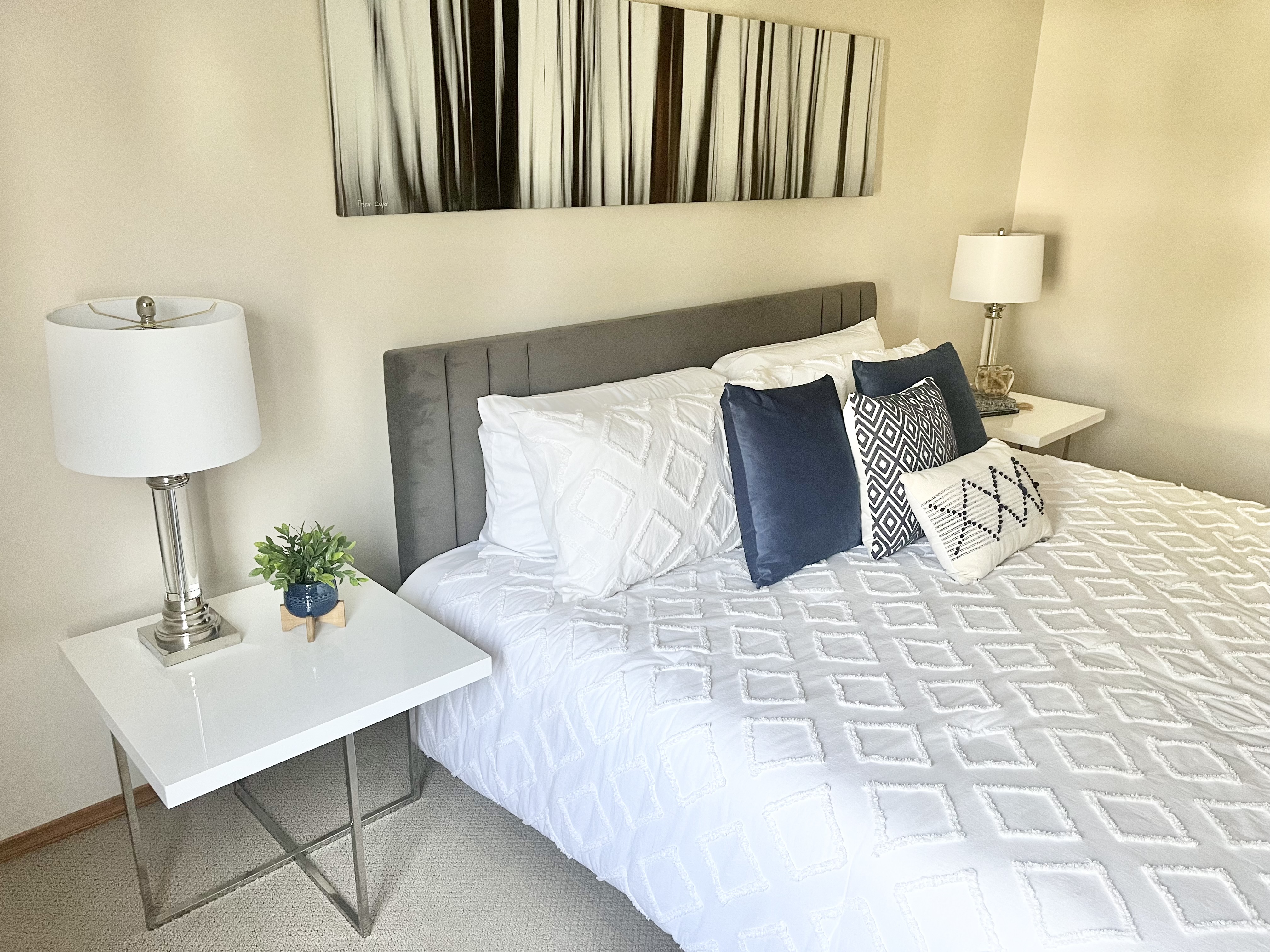 Bright, professionally staged bedroom with a gray upholstered bed, white textured bedding, blue accent pillows, modern bedside tables with lamps, and abstract wall art above the headboard.