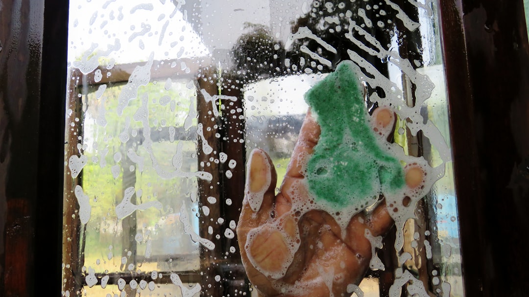 A hand uses a green sponge covered in soap suds to scrub a window, with water droplets and foam spread across the glass.