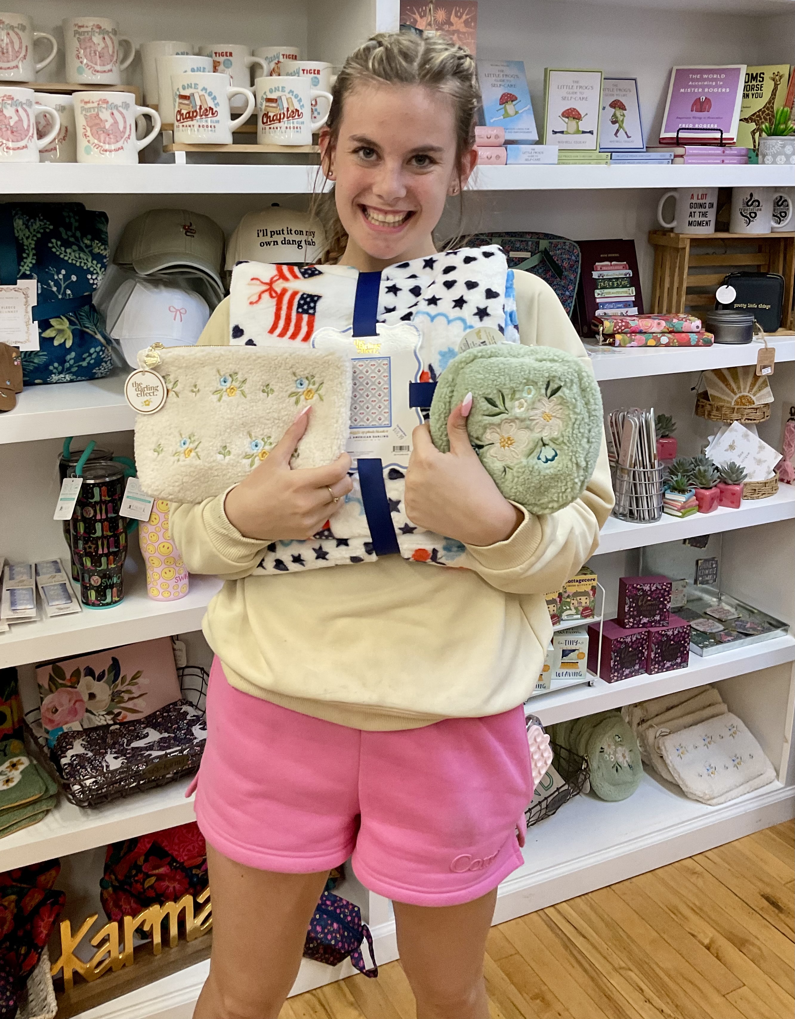 A smiling young woman standing in a boutique gift shop holds a large bundle of retail items. She is carrying a folded plush blanket with an American flag and star pattern, a fluffy cream-colored zipper pouch with embroidered blue flowers, and a small, fuzzy sage green pouch. She wears a beige sweatshirt and pink shorts, with shelves of mugs, hats, and gifts visible behind her.