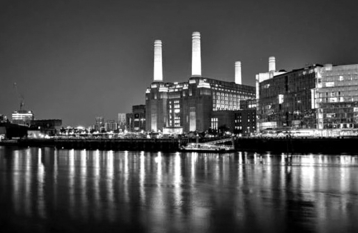 Battersea Power Station lit at night, its four white chimneys rising above the redeveloped riverside buildings, with the lights reflected in the Thames