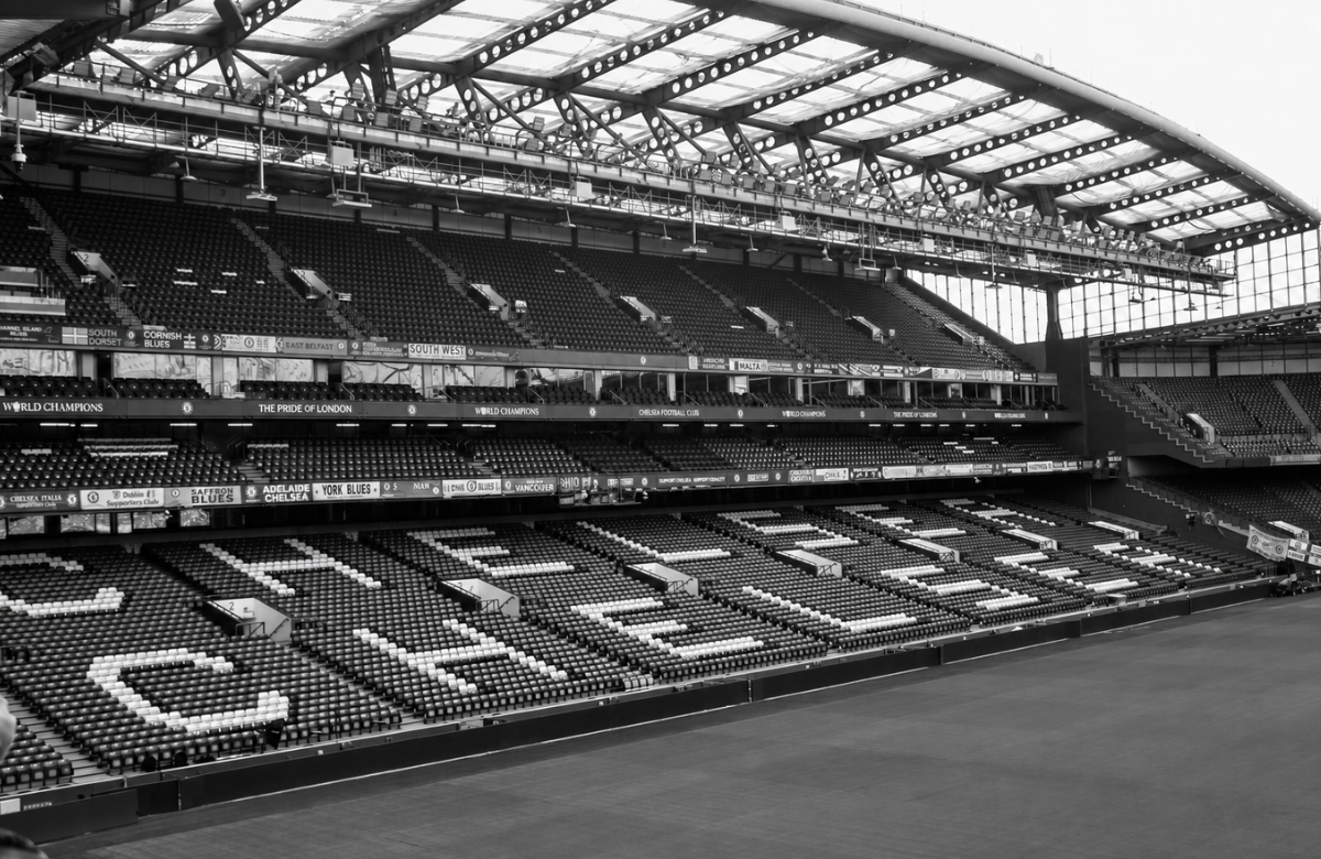 The stands at Stamford Bridge seen from pitch level, empty before a match, with CHELSEA spelled out in white seats and supporters' club banners along the tiers