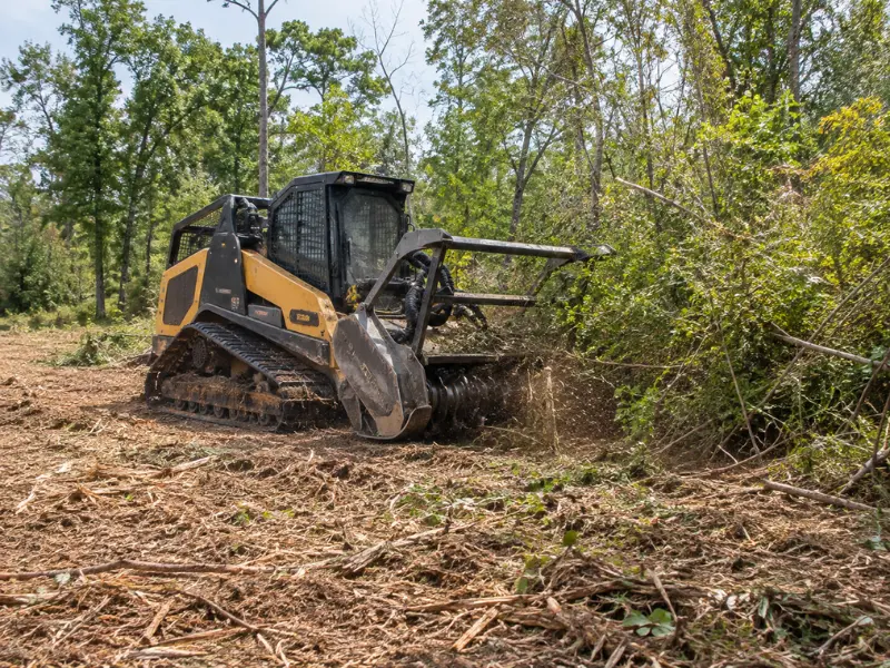 Skid steer with a forestry mulcher attachment grinding through dense brush and small trees during land clearing. Skid steer with a forestry mulcher attachment grinding through dense brush and small trees during land clearing.