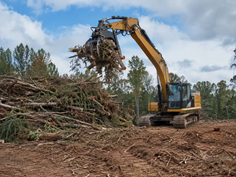 Excavator with a grapple attachment moving cleared trees and brush into a debris pile during land clearing.