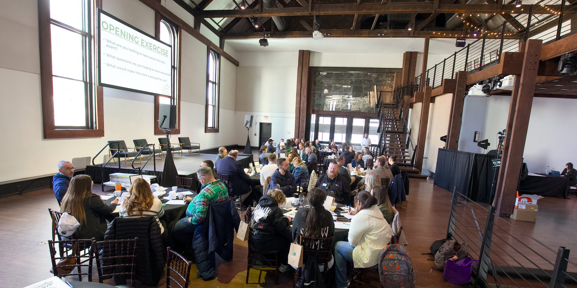 A wide view of the room at the last Kickoff: attendees at round tables working through the opening exercise, with the slide on screen behind them