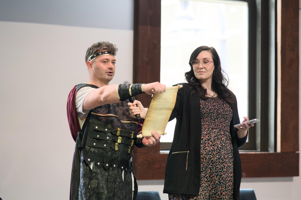 A speaker in costume reading from a scroll on stage beside a host