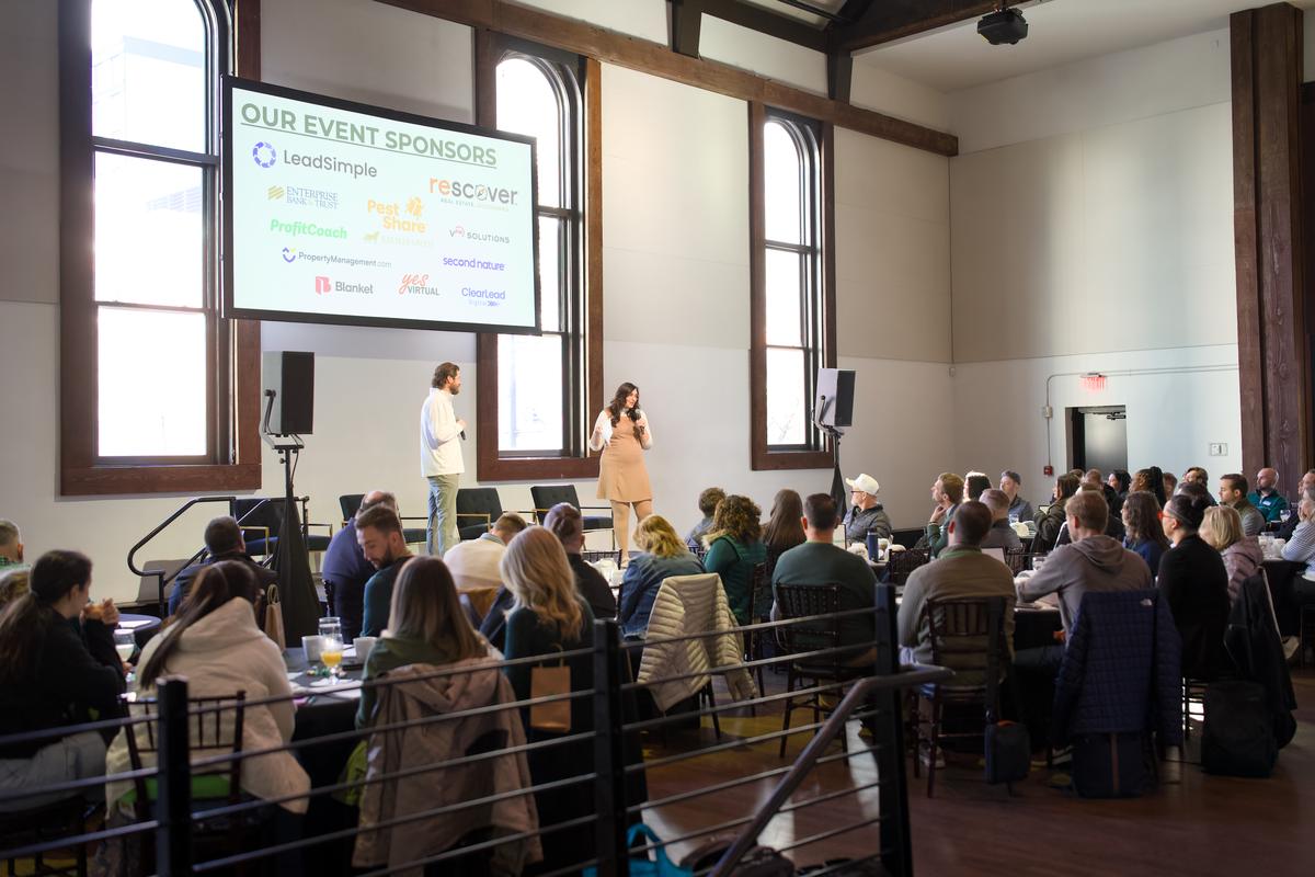 Two hosts presenting from the stage to a full room seated at round tables