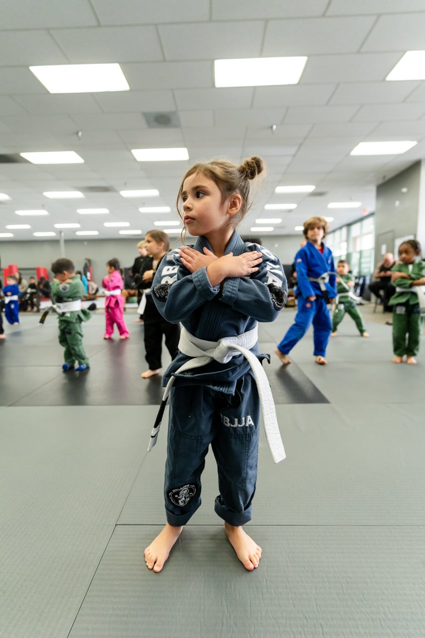 Young boy in a martial arts, walking confidently on mats, wearing a black gi and holding an orange belt..