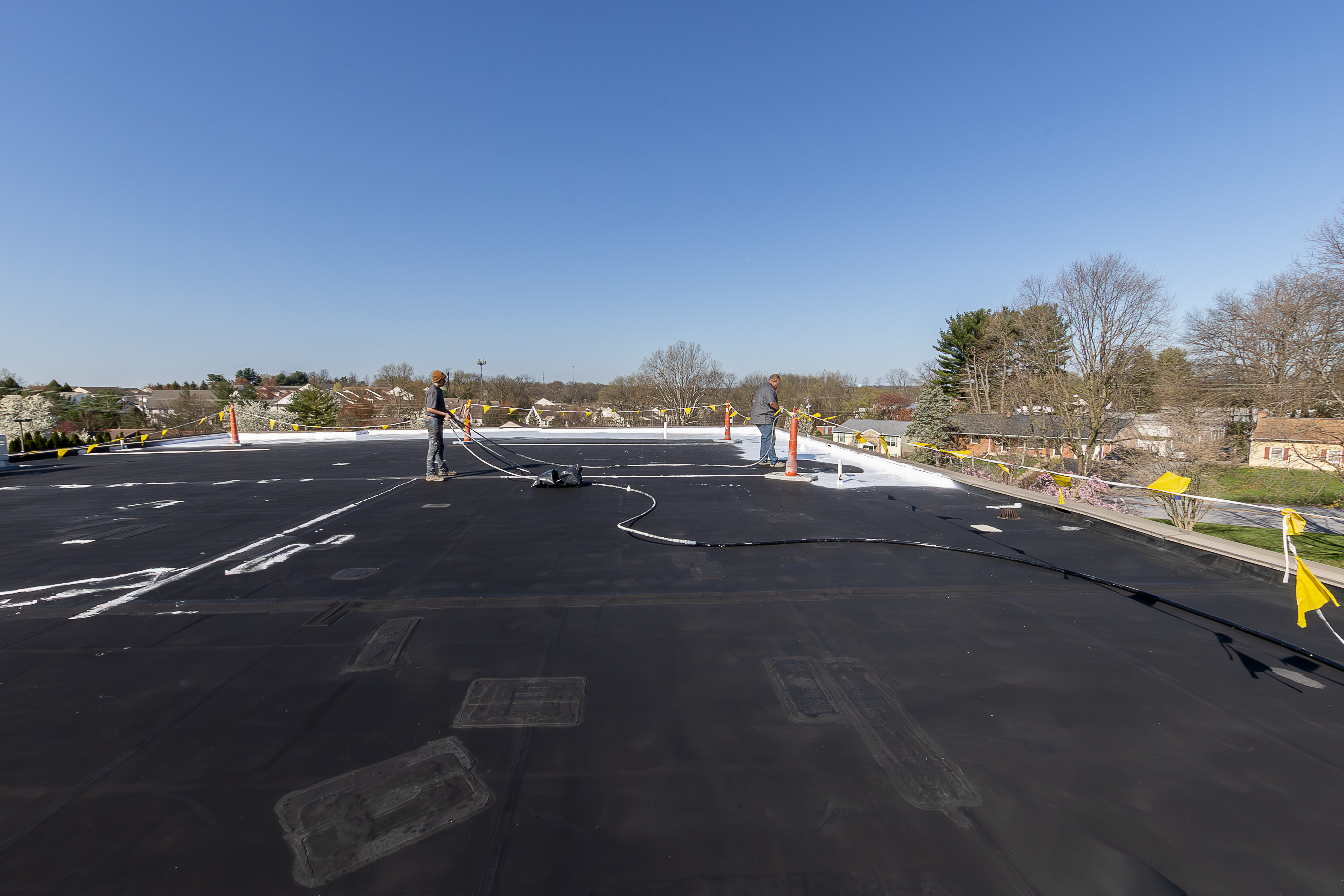 Professional roofers applying protective coating to a large commercial flat roof system on a clear day, demonstrating comprehensive roof restoration services. - Schultz Roofing, Inc Professional roofers applying protective coating to a large commercial flat roof system on a clear day, demonstrating comprehensive roof restoration services. - Schultz Roofing, Inc