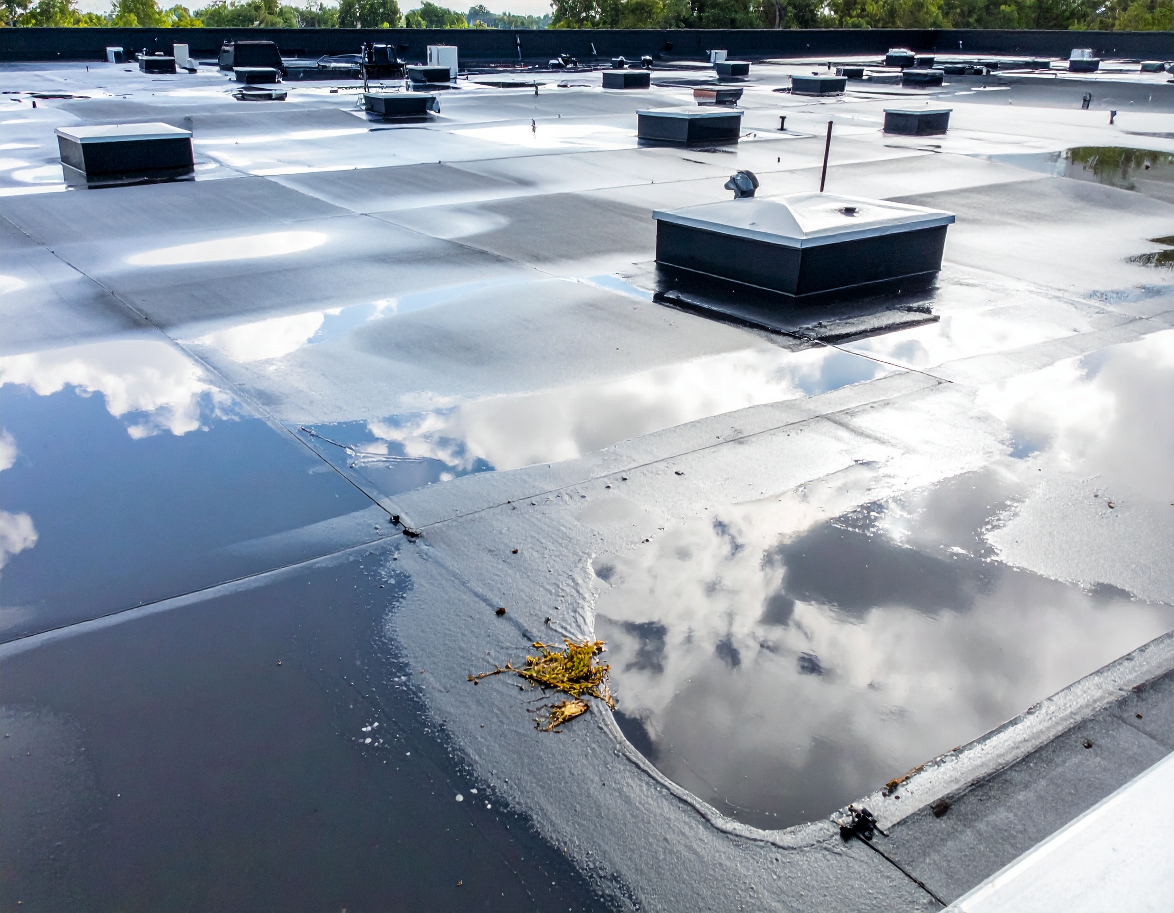 A flat commercial roof showing significant water ponding issues with standing water pools reflecting the sky, along with multiple HVAC units and rooftop equipment throughout the membrane surface. - Schultz Roofing, Inc A flat commercial roof showing significant water ponding issues with standing water pools reflecting the sky, along with multiple HVAC units and rooftop equipment throughout the membrane surface. - Schultz Roofing, Inc