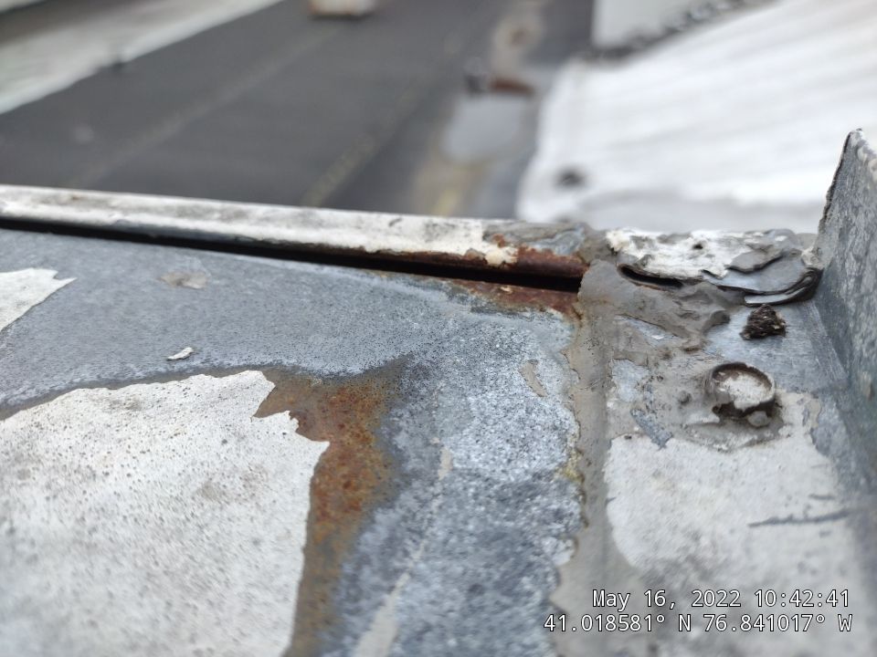 Close-up view of a deteriorating flat roof showing significant rust damage, peeling coatings, and metal corrosion around roof penetrations and seams. - Schultz Roofing, Inc