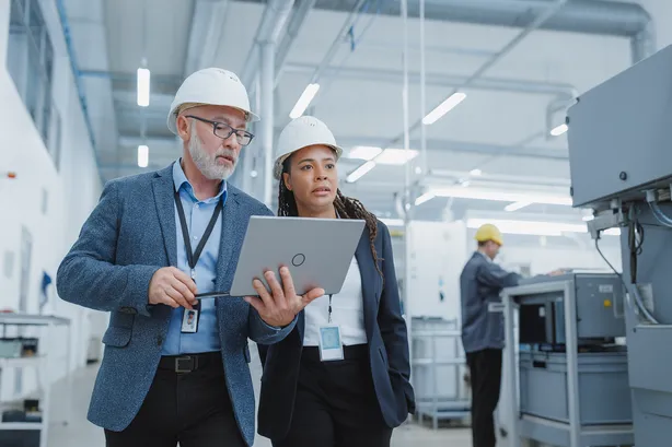 Two engineers wearing safety helmets and formal attire walking through a modern industrial facility, reviewing data on a laptop.