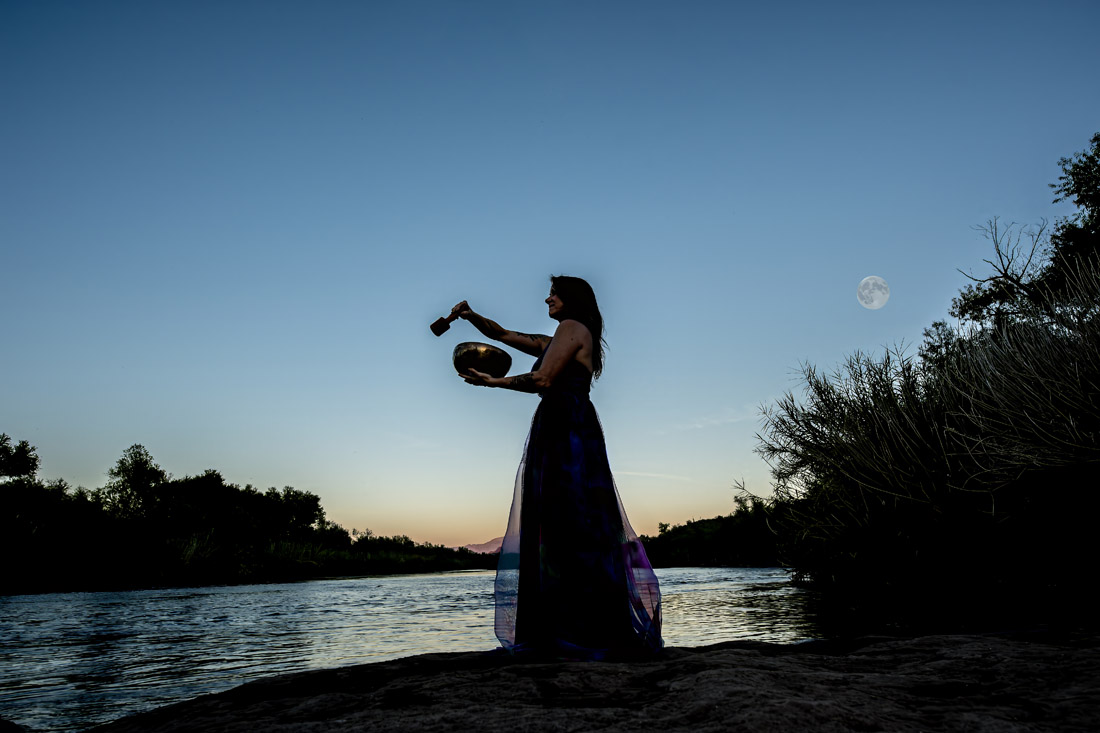 Deb Caron playing a singing bowl at sunset by the river under a full moon