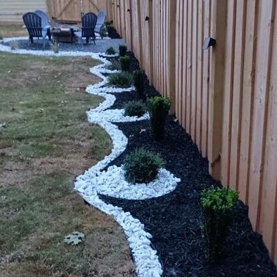 Decorative rock border with mulch and plants along a wooden fence