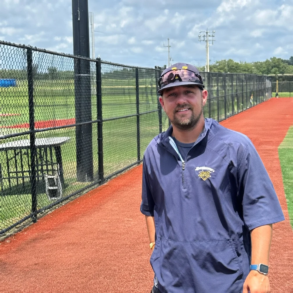 A smiling baseball coach in a navy blue shirt and cap stands on the red dirt track of a baseball field.