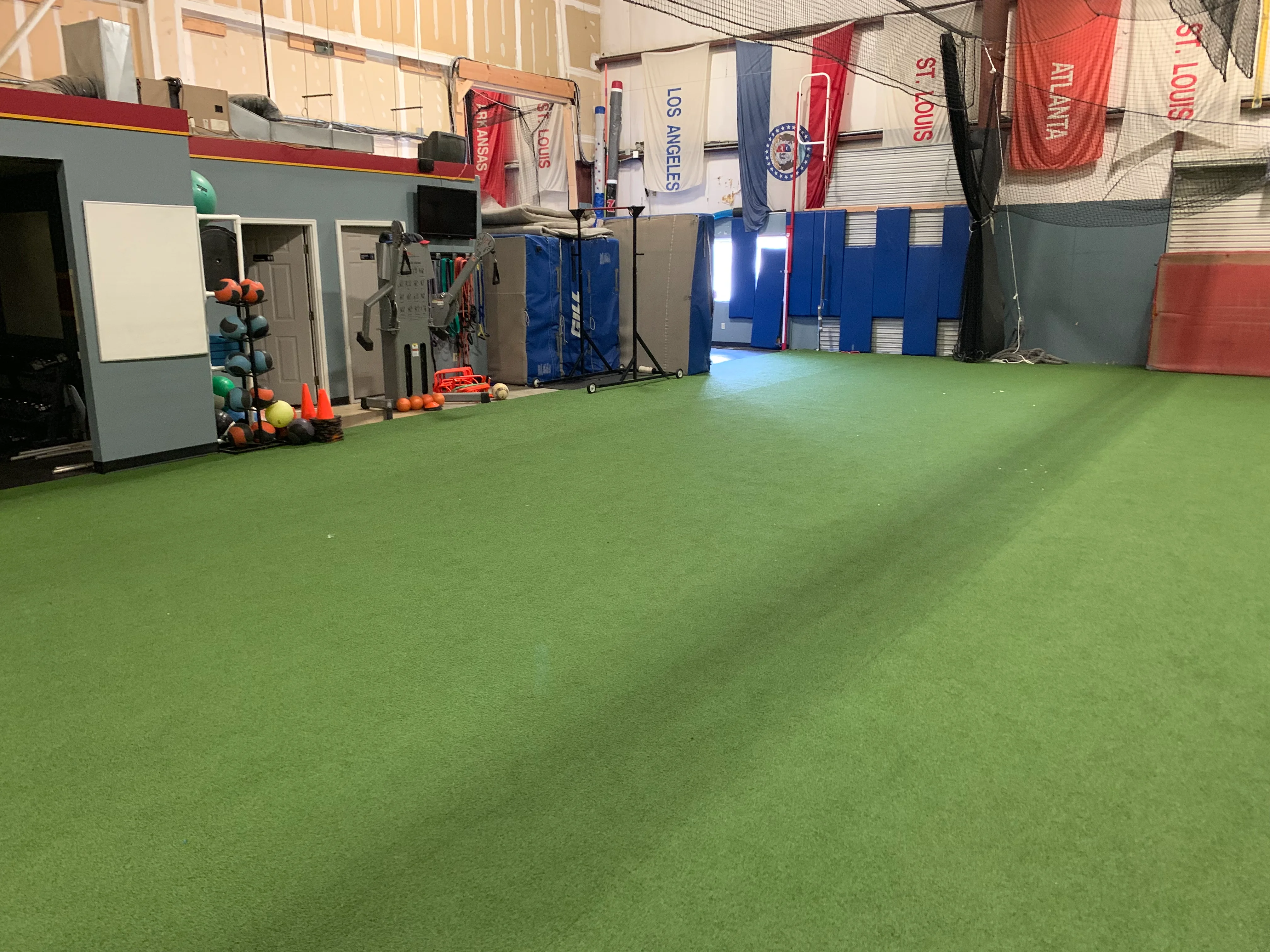 A wide interior shot of an indoor sports training facility with green turf and equipment along the back wall.