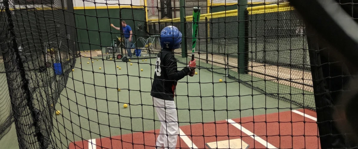 A young baseball player in a helmet and uniform stands at home plate, holding a bat, in an indoor batting cage.