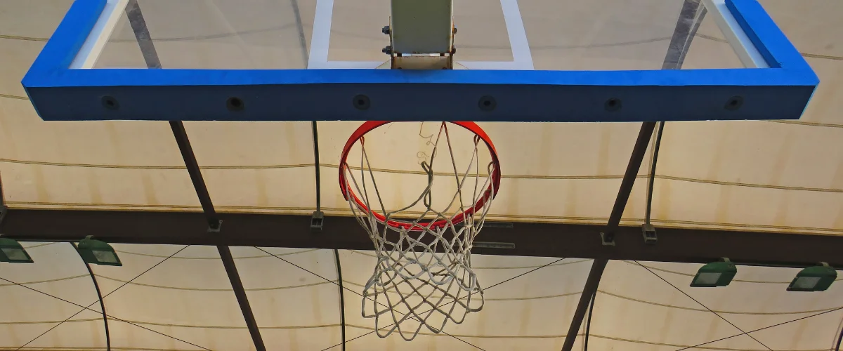 A close-up, bottom-up view of a red and white basketball hoop with a clear backboard under a tented ceiling.