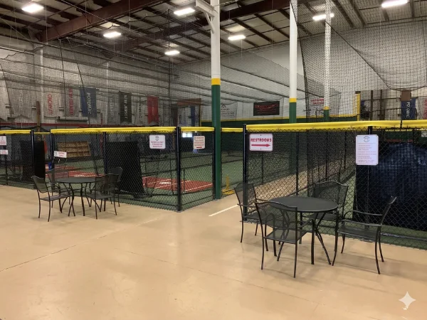 An indoor batting cage facility with chain link fencing, green turf, and two small tables with chairs.