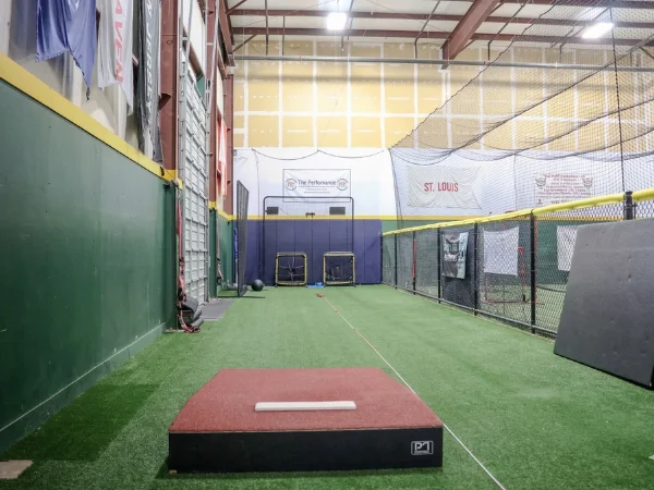An empty indoor baseball training facility with green turf and a red pitcher's mound in the foreground.
