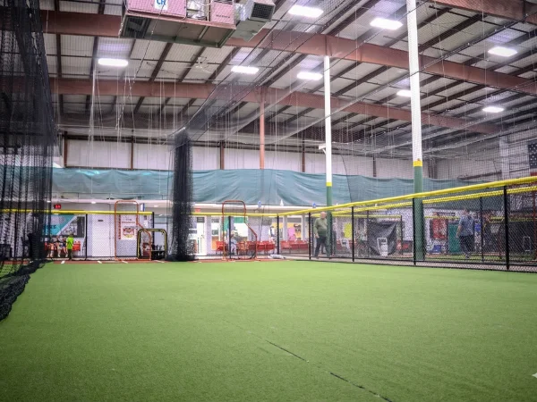 Wide shot of an indoor baseball training facility with green turf and multiple batting cages.