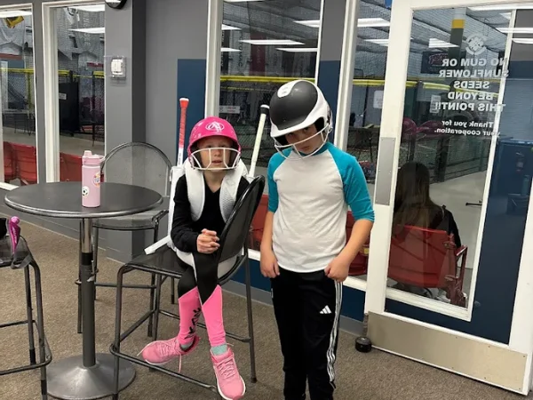 Two children wearing baseball batting helmets and athletic gear stand near a high-top table beside batting cages.