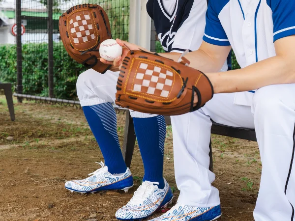 Two baseball players in uniform sitting on a bench, one holding a ball and wearing a brown glove.