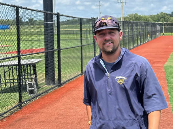 Instructor standing by a fenced baseball field wearing a cap and training jacket
