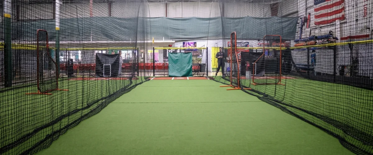 Wide shot of an empty indoor batting cage facility with green turf and protective netting.