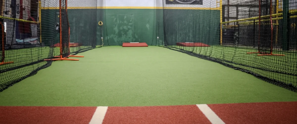 An empty indoor batting cage with green turf, dark netting, pitching mounds, and a red batter's box.