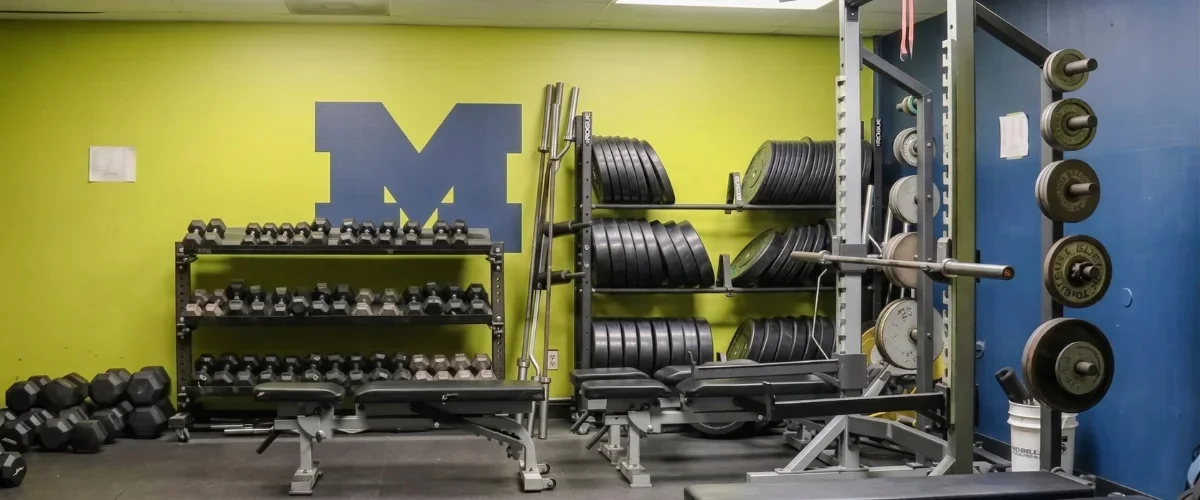 A gym with weight racks, dumbbells, benches, and a large blue M logo on a yellow wall.