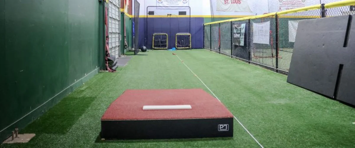 A baseball pitching mound with artificial turf is positioned at the center of an indoor batting cage.