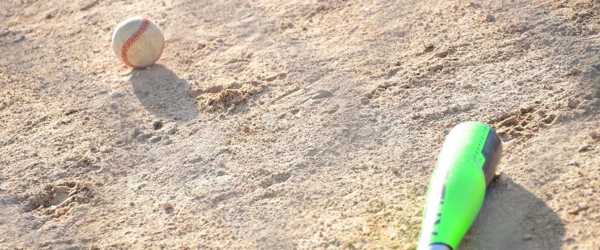 A baseball with red stitching and a green composite bat resting on sandy dirt ground in bright sunlight.