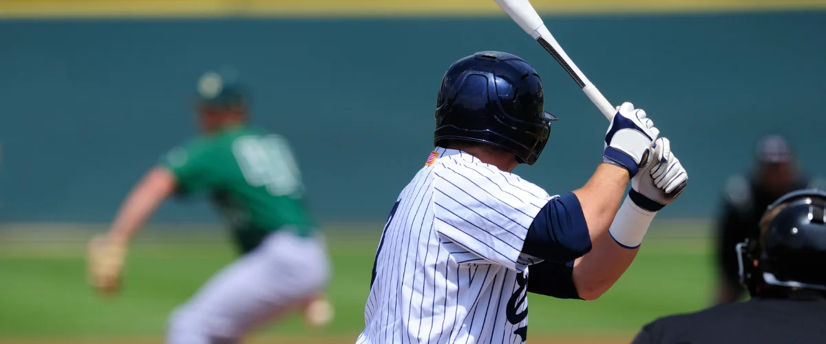 A baseball batter in a striped uniform and navy helmet waits with a bat at home plate, with a pitcher in green in the background. A baseball batter in a striped uniform and navy helmet waits with a bat at home plate, with a pitcher in green in the background.