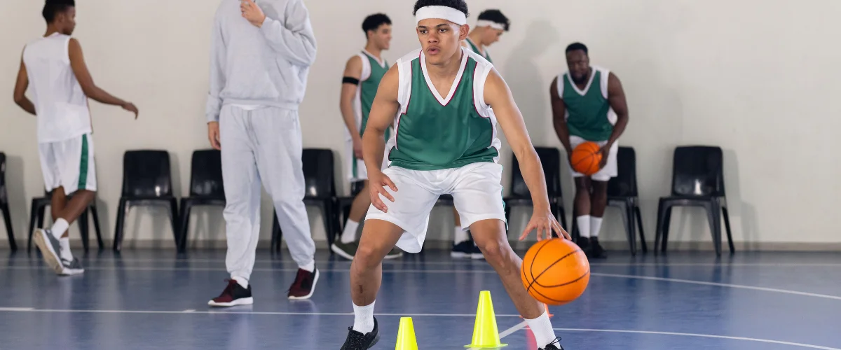 A young male basketball player in a green jersey dribbling a ball between yellow cones during a drill. A young male basketball player in a green jersey dribbling a ball between yellow cones during a drill.