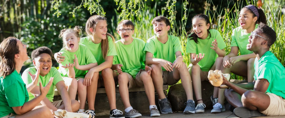 A diverse group of happy children in bright green shirts laughing and sitting together outdoors with an adult. A diverse group of happy children in bright green shirts laughing and sitting together outdoors with an adult.