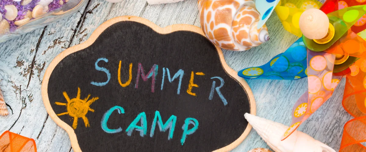 A chalkboard sign saying "SUMMER CAMP" with a sun drawing, surrounded by a pinwheel, shells, and colorful ribbons. A chalkboard sign saying "SUMMER CAMP" with a sun drawing, surrounded by a pinwheel, shells, and colorful ribbons.