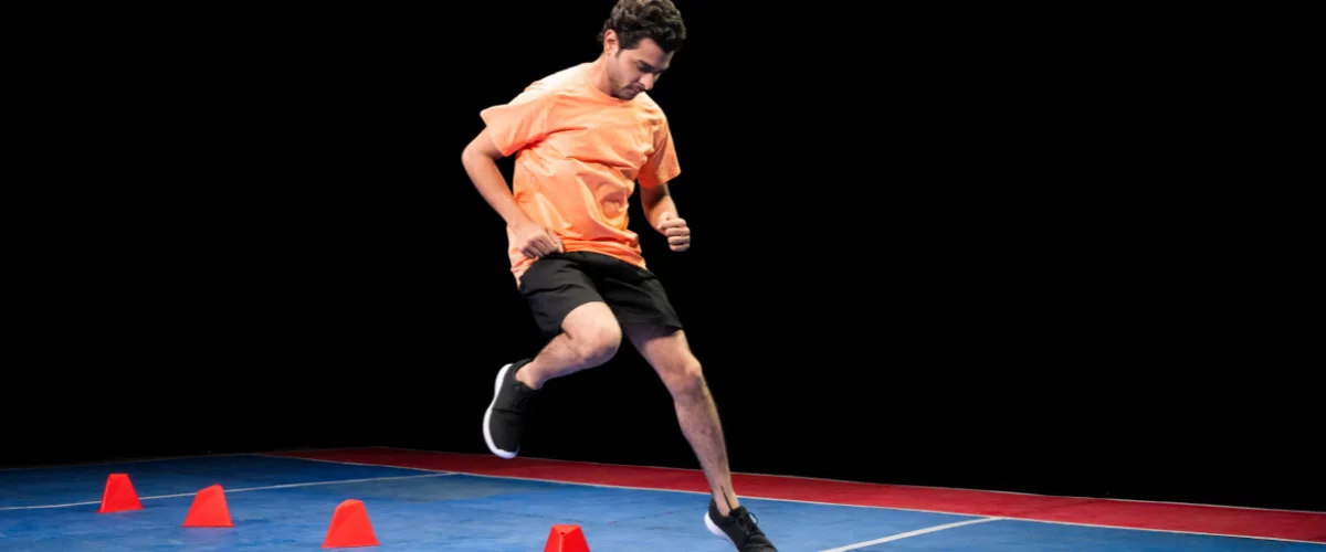 A man in an orange shirt and black shorts performs agility drills, running with three orange cones on a blue and red court.